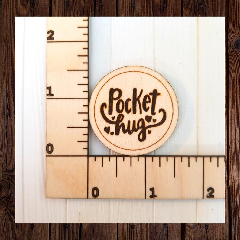 May include: A round wooden tag with the text "Pocket hug" in black cursive lettering with two small hearts above the word "hug".