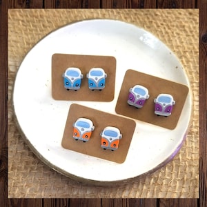 May include: A set of six pairs of earrings featuring miniature white Volkswagen bus designs. Each pair has a different color: blue, orange, purple, and white. The earrings are displayed on a white plate with brown cardstock backing.