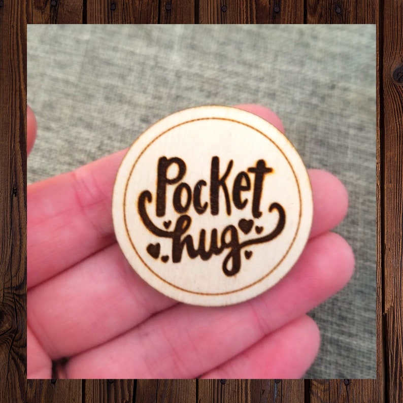 May include: A round wooden disc with the words "Pocket hug" engraved in a cursive font. The words are surrounded by three small hearts.