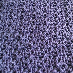 May include: A purple crocheted fabric featuring a repeating openwork stitch pattern. The textile is composed of interconnected yarn loops that create a textured, semi-sheer surface with a consistent, dense arrangement of small, uniform, shell-like clusters.