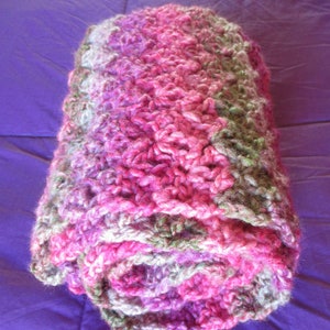 May include: A pink, green, and white crocheted blanket rolled up. The blanket is made of soft yarn and has a textured, fluffy look.