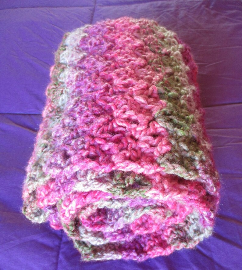 May include: A pink, green, and white crocheted blanket rolled up. The blanket is made of soft yarn and has a textured, fluffy look.