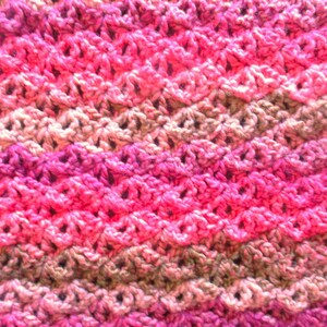 May include: A crocheted scarf with a pink, green, and beige color pattern. The scarf is made with a textured stitch that creates a wave-like design.