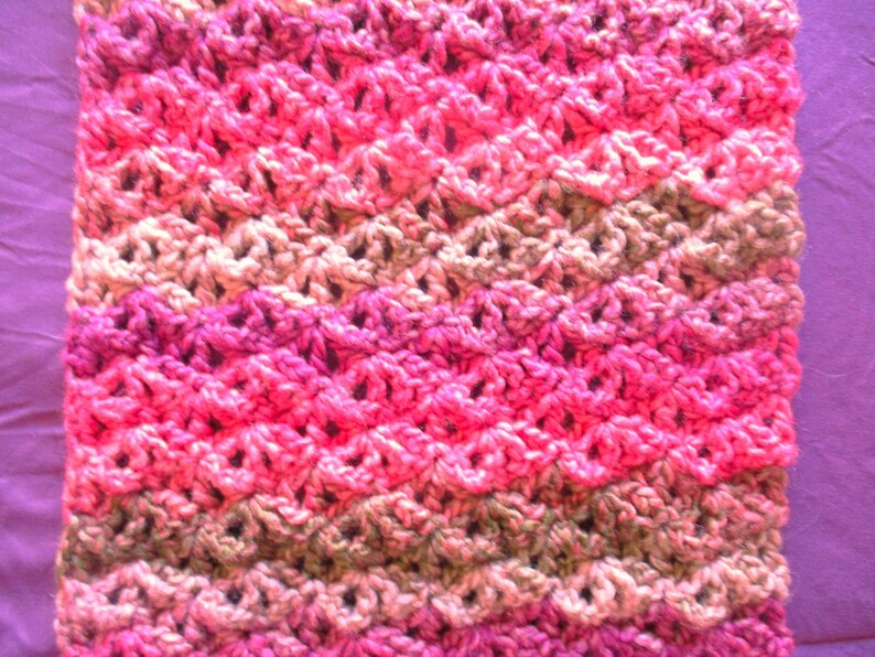 May include: A crocheted scarf with a pink, green, and beige color pattern. The scarf is made with a textured stitch that creates a wave-like design.
