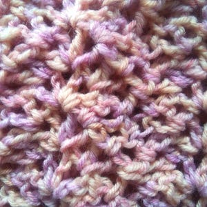 Pode incluir: Close-up de um t&ecirc;xtil tricotado &agrave; m&atilde;o com uma trama texturizada e aberta. O fio &eacute; uma mistura de cores, incluindo p&ecirc;ssego, lavanda e creme, criando uma apar&ecirc;ncia suave e misturada. O tecido de malha tem uma estrutura densa, mas arejada.