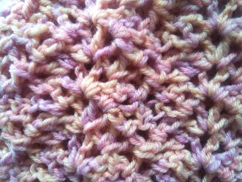 Pode incluir: Close-up de um t&ecirc;xtil tricotado &agrave; m&atilde;o com uma trama texturizada e aberta. O fio &eacute; uma mistura de cores, incluindo p&ecirc;ssego, lavanda e creme, criando uma apar&ecirc;ncia suave e misturada. O tecido de malha tem uma estrutura densa, mas arejada.