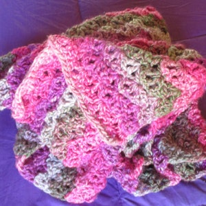 May include: A crocheted blanket in shades of pink, purple, and green. The blanket is made with a textured stitch and has a ruffled edge.