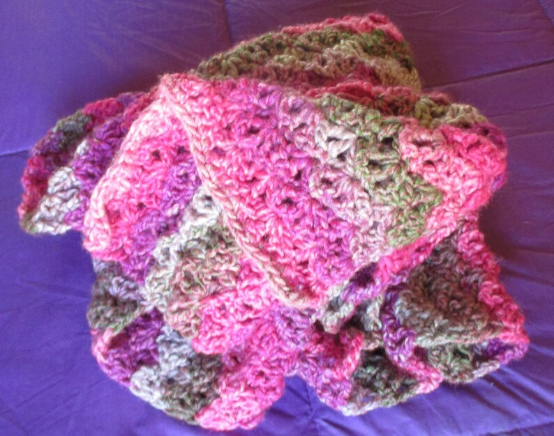 May include: A crocheted blanket in shades of pink, purple, and green. The blanket is made with a textured stitch and has a ruffled edge.
