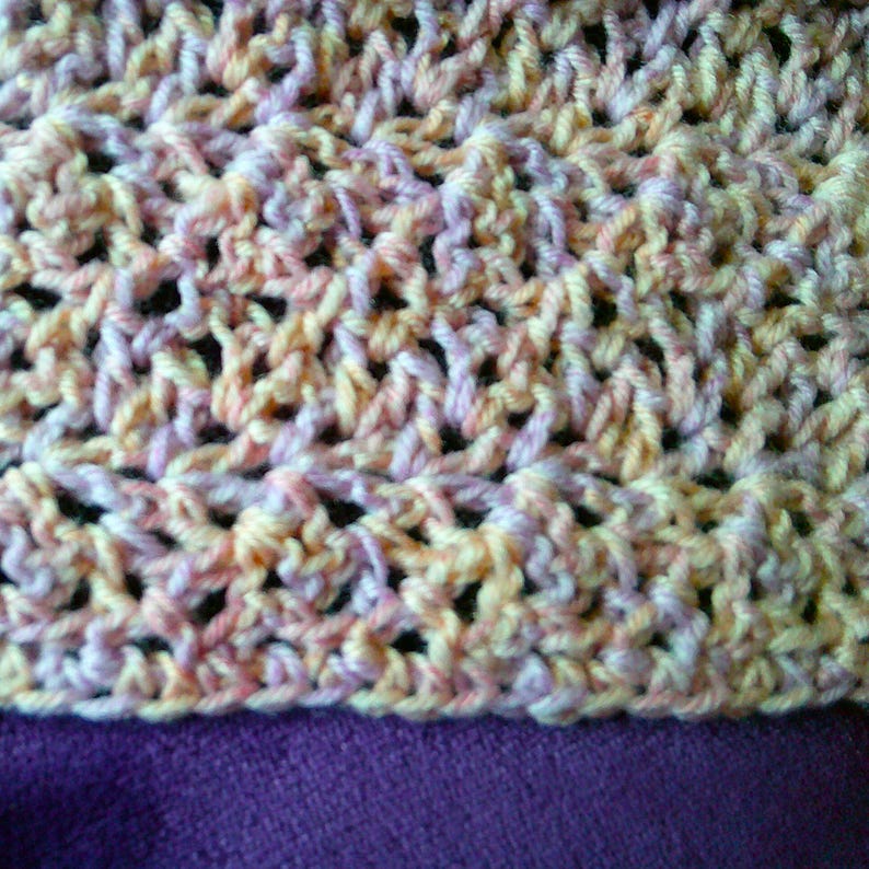 Pode incluir: Close-up de um tecido de croch&ecirc; em tons de amarelo, p&ecirc;ssego e lavanda. O padr&atilde;o texturizado &eacute; vis&iacute;vel, com um fundo roxo. O fio parece macio e os pontos s&atilde;o bem tecidos, criando um tecido denso e padronizado.