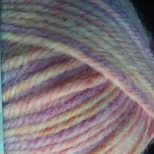 Pode incluir: Close-up de um novelo de l&atilde; em tons de lavanda, rosa, p&ecirc;ssego e creme. A l&atilde; &eacute; espessa e texturizada, com fios vis&iacute;veis. As cores misturam-se num padr&atilde;o suave e variado, ideal para projetos de tric&ocirc; ou croch&ecirc;.