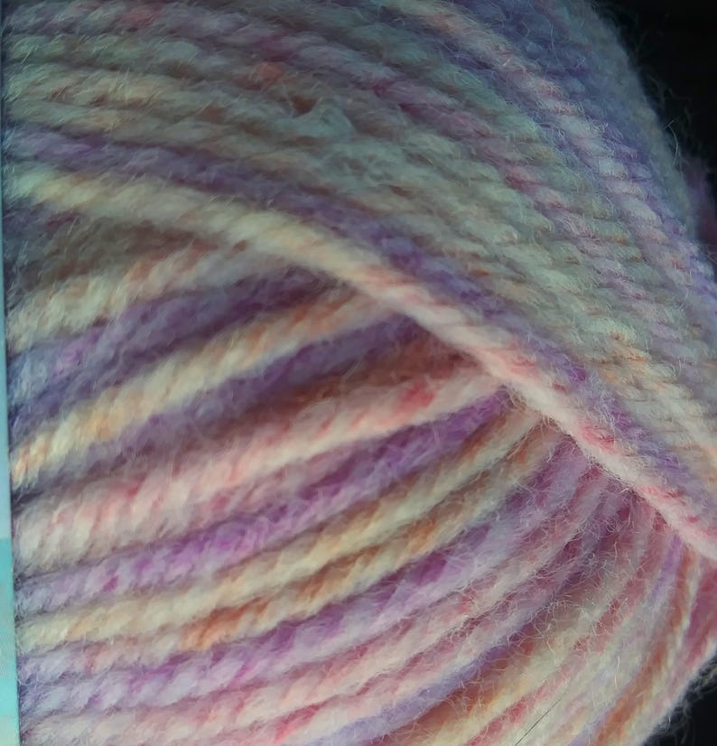 Pode incluir: Close-up de um novelo de l&atilde; em tons de lavanda, rosa, p&ecirc;ssego e creme. A l&atilde; &eacute; espessa e texturizada, com fios vis&iacute;veis. As cores misturam-se num padr&atilde;o suave e variado, ideal para projetos de tric&ocirc; ou croch&ecirc;.