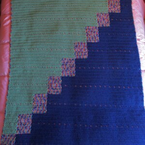 May include: A crocheted blanket with a diagonal pattern of blue and green squares. The squares are outlined in a multi-colored yarn.