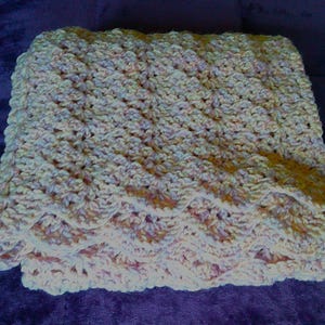 Pode incluir: Um cobertor de croch&ecirc; feito &agrave; m&atilde;o, dobrado, em cores pastel suaves. O cobertor apresenta um padr&atilde;o de ponto texturizado no corpo principal e uma borda ondulada. A paleta de cores inclui tons de creme, rosa e amarelo p&aacute;lido, criando uma est&eacute;tica acolhedora e convidativa.