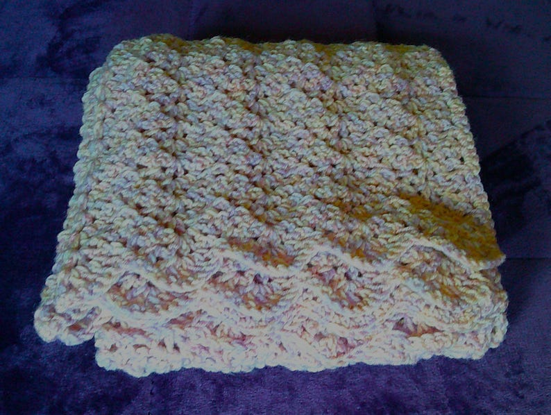 Pode incluir: Um cobertor de croch&ecirc; feito &agrave; m&atilde;o, dobrado, em cores pastel suaves. O cobertor apresenta um padr&atilde;o de ponto texturizado no corpo principal e uma borda ondulada. A paleta de cores inclui tons de creme, rosa e amarelo p&aacute;lido, criando uma est&eacute;tica acolhedora e convidativa.