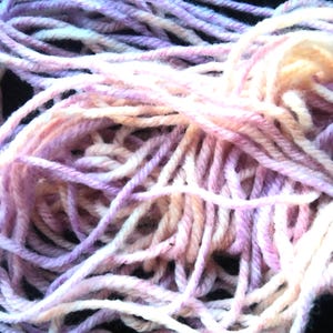 Pode incluir: Close-up de um novelo de l&atilde; em tons de lavanda, rosa e creme. A l&atilde; &eacute; enrolada soltamente, mostrando os fios individuais. Ideal para tric&ocirc;, croch&ecirc; ou outros projetos de artes t&ecirc;xteis.