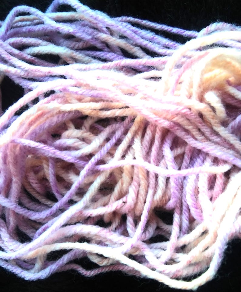 Pode incluir: Close-up de um novelo de l&atilde; em tons de lavanda, rosa e creme. A l&atilde; &eacute; enrolada soltamente, mostrando os fios individuais. Ideal para tric&ocirc;, croch&ecirc; ou outros projetos de artes t&ecirc;xteis.