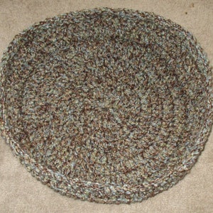 May include: A round crocheted rug in shades of brown, green, and blue. The rug has a textured, woven appearance.