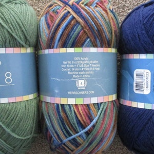May include: Three balls of worsted weight yarn. The first ball is green, the second is a multi-colored mix of brown, blue, and green, and the third is navy blue. The labels on the balls of yarn are from Herrschners and include the yarn weight, fiber content, and care instructions.