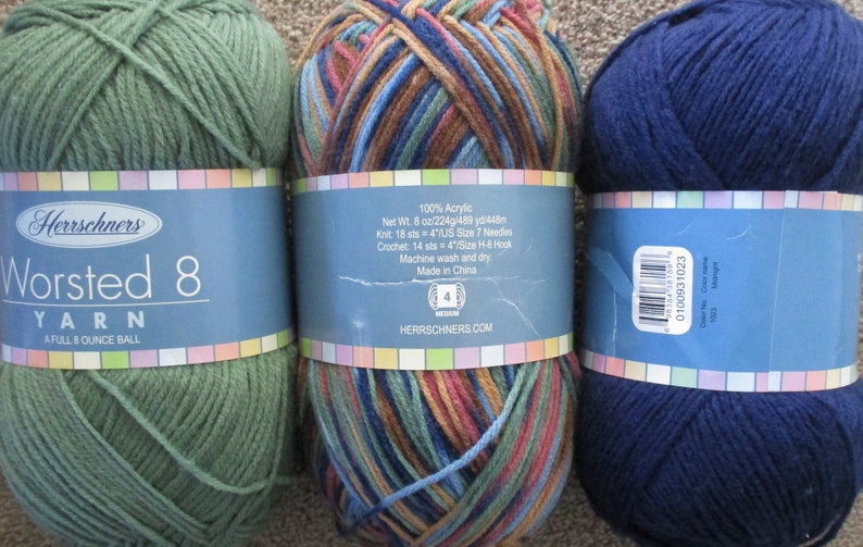 May include: Three balls of worsted weight yarn. The first ball is green, the second is a multi-colored mix of brown, blue, and green, and the third is navy blue. The labels on the balls of yarn are from Herrschners and include the yarn weight, fiber content, and care instructions.