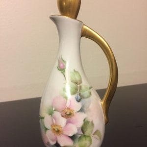 May include: A white porcelain pitcher with a gold handle and a gold top. The pitcher is decorated with hand-painted pink roses and green leaves.