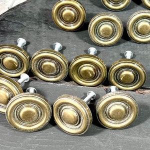 May include: A collection of twelve round cabinet knobs featuring a metallic antique brass finish. Each knob has a concentric circular design with a raised center point and a silver-toned metal screw post extending from the back side.