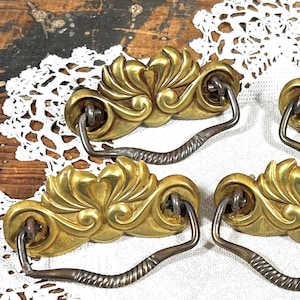 May include: A set of four brass drawer pulls featuring ornate, fan-shaped backplates with swirling, leaf-like details and attached swinging, twisted metal bail handles. Each piece displays a polished gold-tone finish with dark metal accents.