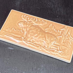 May include: A rectangular, tan-colored ceramic tile featuring a raised relief of a moose in a forest setting. The moose has large antlers and is surrounded by trees. The tile has a glossy finish and is likely for decorative use.