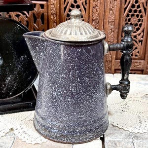 May include: A vintage speckled enamelware coffee pot in dark blue with a silver lid and black handle. The pot is displayed on a stone surface, with a black scale and red decorative apples in the background.