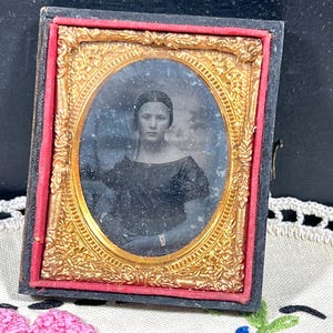 May include: Antique framed portrait featuring a woman in a dark dress. The oval portrait is set within a gold-colored ornate frame, surrounded by a red border, and encased in a black frame. The portrait is displayed on a white embroidered surface.