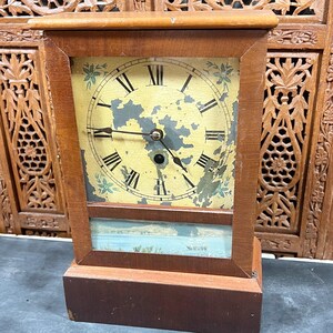 May include: A vintage wooden clock with a rectangular frame. The clock face has Roman numerals and decorative floral accents. The clock's face is yellow with some paint peeling. The bottom section of the clock has a landscape painting.