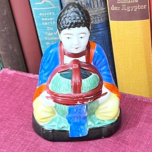May include: A vintage porcelain figurine of an Asian figure holding a basket. The figurine is dressed in a blue robe with orange and yellow accents. The basket is green with a red handle. The figurine is set against a backdrop of antique books.