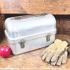 May include: A silver metal toolbox with a brown handle and clasps sits on a wooden surface next to a pair of work gloves, a red apple, and a leather tool belt with tools.