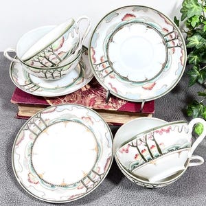 May include: A set of white porcelain teacups and saucers with a tree design. The cups and saucers have a delicate, detailed pattern in shades of green, red, and brown. The set is arranged on a dark red book and a gray surface.