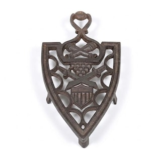 May include: A cast iron trivet with a shield shape and a decorative handle. The trivet features crossed swords, grapes, and a crest design. The color is a dark brown.