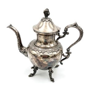 May include: A silver-plated teapot featuring an ornate handle, a curved spout, and three decorative feet. The lid is topped with a grape cluster finial, and the body is embellished with embossed grape vine patterns around the neck and base.