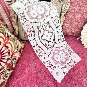 May include: A white rectangular table runner featuring intricate cutwork embroidery with floral motifs and scalloped edges. The lace-like fabric displays detailed needlework patterns of blossoms and leaves throughout the entire length of the piece.