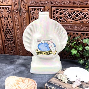 May include: A pearlescent, shell-shaped ceramic bottle featuring a graphic of the state of Florida superimposed over a blue globe. A yellow banner at the bottom displays the text, Sea Shell Headquarters of the World, in black lettering.