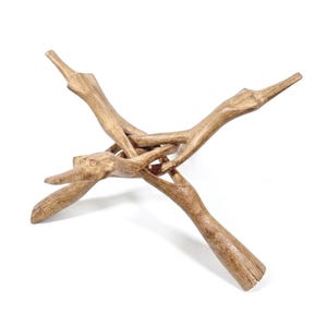 May include: A handcrafted wooden display stand with a unique, intertwined branch design. The stand is made of light brown wood and features four intersecting branches, creating a stable base for displaying small objects. The stand is designed to hold a small bowl or decorative item.