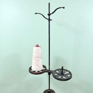 May include: An antique black metal thread stand with a spool of white thread. The stand has a decorative design with two circular platforms and a curved arm for holding the thread. The base is round and has the number "700" on it.