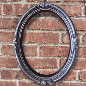 May include: An oval-shaped picture frame with a dark metallic finish. The frame features ornate, raised floral-style carvings at the top, bottom, and both sides. It has a smooth, rounded inner border and a hanging hook attached to the top center.