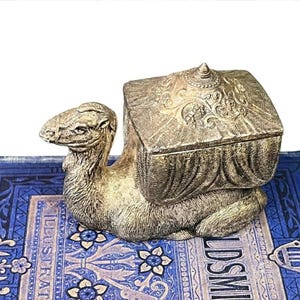 May include: A detailed, silver-colored camel figurine with a decorative box on its back. The camel is lying down, and the box has ornate floral patterns. The figurine is placed on a blue and white patterned surface.