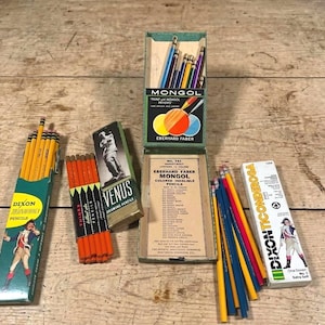 May include: A collection of vintage pencil boxes including a green Dixon Ticonderoga box, a Venus drawing pencil box, an open Eberhard Faber Mongol box with colored pencils, and a white Dixon Ticonderoga box, accompanied by loose colored pencils.