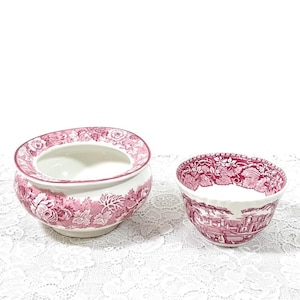 May include: Two ceramic bowls with a pink floral pattern. The larger bowl has a wide, open top and a detailed floral design around the rim and base. The smaller bowl features a landscape scene. Both bowls have a white interior.