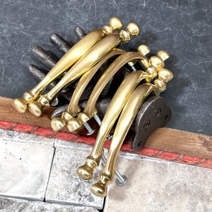 May include: A collection of six brass-colored cabinet pulls. Each pull has a curved handle and decorative end pieces. The pulls are arranged on a dark surface, showcasing their polished finish and classic design. Screws are attached to the pulls.