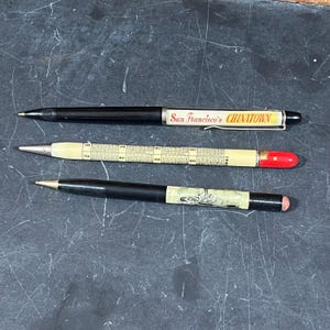 May include: Three vintage pens are displayed against a dark background. One black pen has "San Francisco's Chinatown" printed on it. Another pen is cream-colored with a calendar and a red tip. The third pen is black with a decorative design.