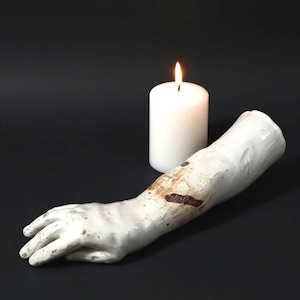 May include: A white ceramic arm sculpture with brown accents, resting on a dark surface. A lit white candle is positioned behind the arm. The sculpture has a textured surface and detailed hand.