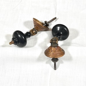 May include: Two drawer pulls featuring smooth, round black knobs attached to ornate, circular brass backplates with intricate embossed patterns. Each pull includes a threaded metal bolt and a hexagonal nut for secure installation on furniture.