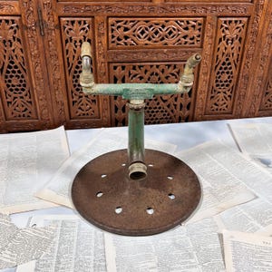 May include: A vintage green metal lawn sprinkler with a circular, rusted base featuring several drainage holes. The central vertical pipe connects to a horizontal bar with two angled nozzles at the ends. The text "CAMPBELL" is embossed on the center.
