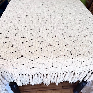 May include: Cream-colored, open-weave tablecloth with a geometric cube pattern. The tablecloth has a fringed edge and is draped over a dark wooden table. The intricate design adds a touch of vintage charm to any dining setting.