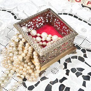 May include: A vintage silver-toned jewelry box with an ornate filigree design, lined with red velvet. The box is filled with strands of faux pearls in varying sizes and shades of white and cream. The pearls cascade over the sides.